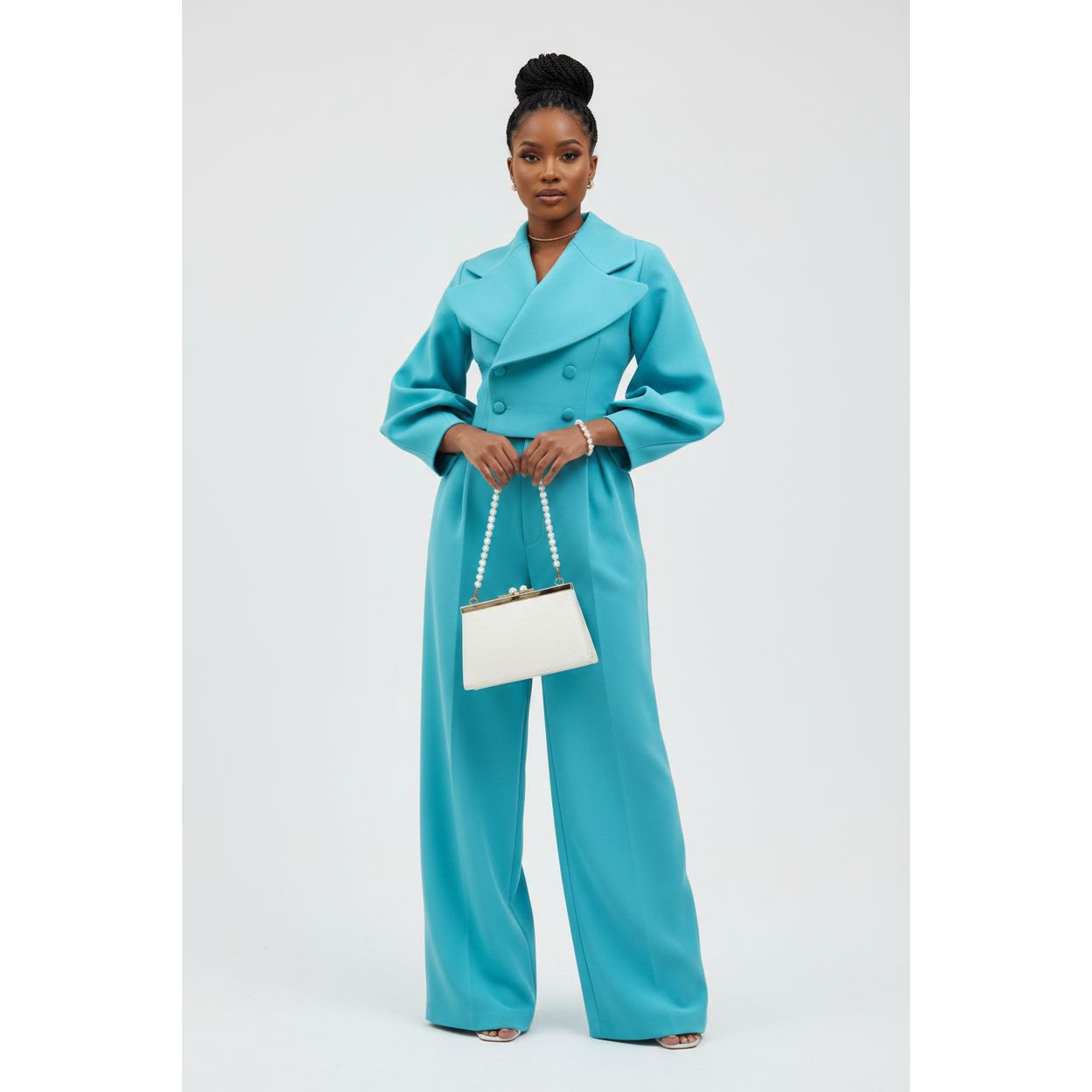 Woman wearing a turquoise coat and pants holding a white handbag on a light gray background
