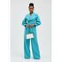 Woman wearing a turquoise coat and pants holding a white handbag on a light gray background