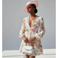 Woman wearing a floral dress and pink hat with a white handbag against a gray background