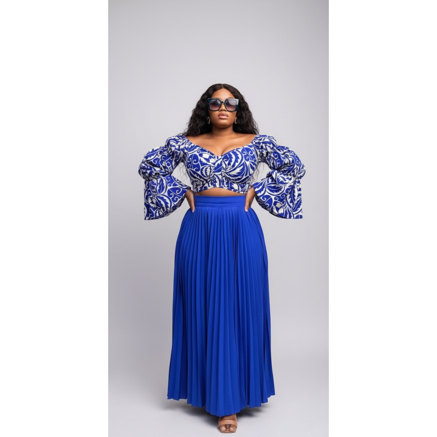 Woman wearing a blue and white patterned top with a pleated blue skirt on a gray background