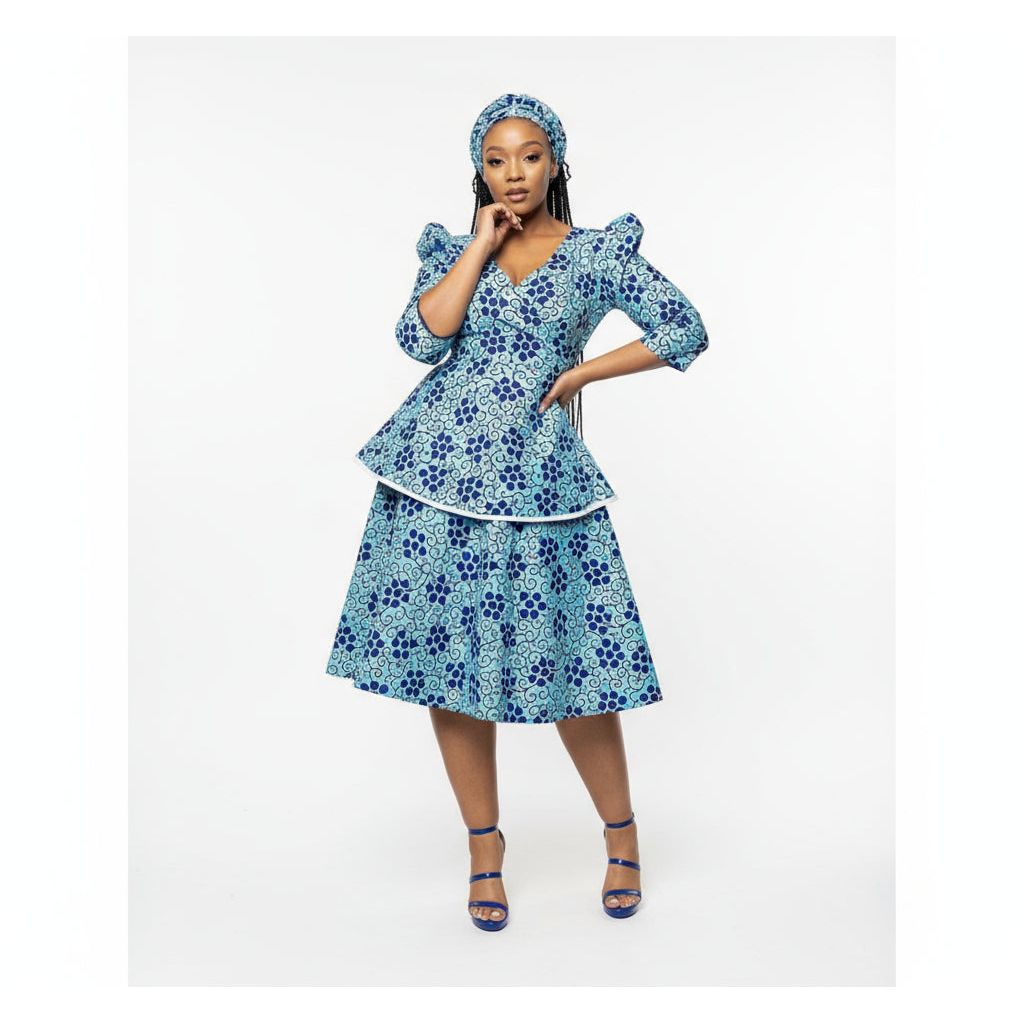 A woman wearing a blue floral African print pleated midi dress with a V-neck and 3/4 sleeves, paired with a matching headwrap. She is standing in a room with elegant decor.