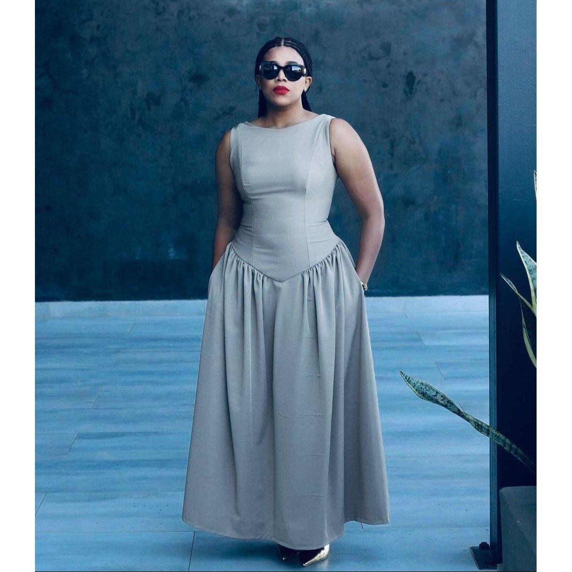 A woman wearing a sleeveless corset pleated dress in a beige color, standing in a sunny location with a plant background.