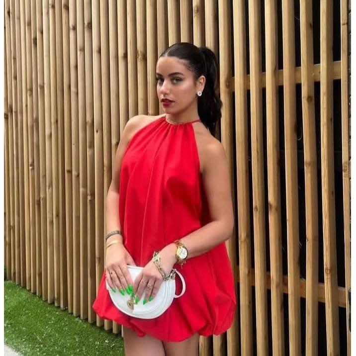 A woman wearing a red sleeveless chiffon balloon bubble dress with a halter neck, showcasing a white and green handbag and gold accessories.