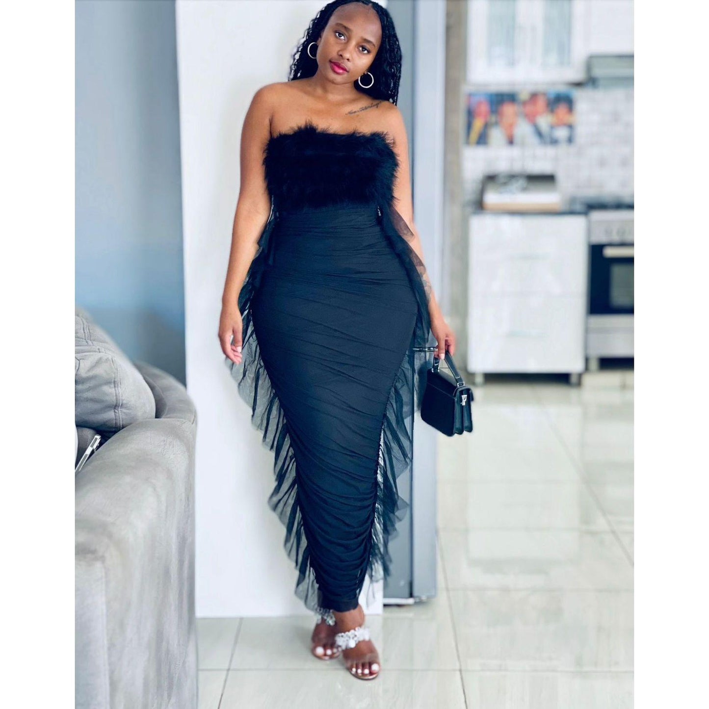 A woman wearing a black strapless maxi dress with fur detailing on the top and fringe along the skirt, standing in a home environment.