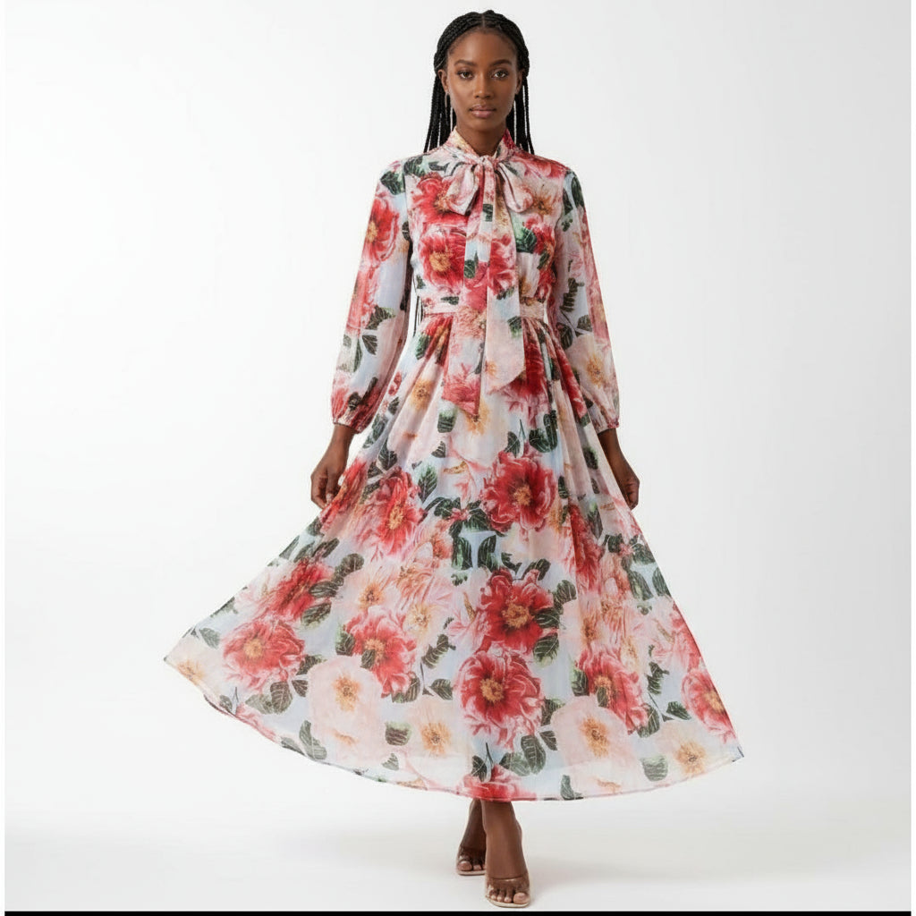 A long floral chiffon dress displayed on a surface, with prominent red flowers and green leaves pattern, and a bow at the waist.