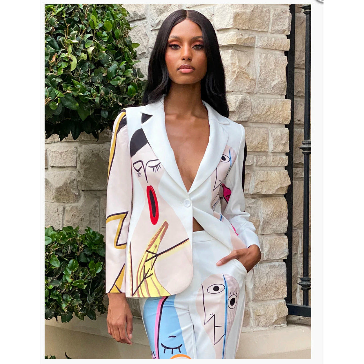 A woman wearing a cartoon print twin set with vivid colors, consisting of a blazer and pants, standing near a brick wall.